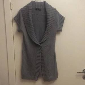 Grå stickad kofta från Mexx - Säljer en supermysig grå stickad kofta från Mexx gjord i bomull. Den har korta ärmar och en bred ribbad krage som ger en extra touch av stil. Perfekt för lager-på-lager under höst och vinter. Koftan är i bra skick och redo för nya äventyr! 🌟