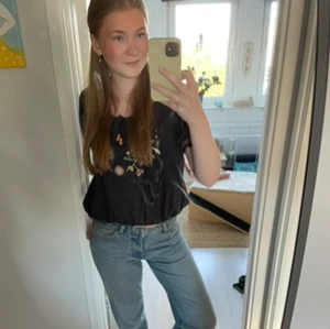 Svart blus med broderade blommor - Säljer en superfin svart blus med färgglada broderade blommor på bröstet. Blusen har korta ärmar och en söt volangdetalj längst ner. Perfekt för både vardag och fest! 🌸