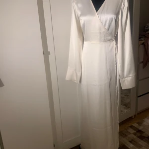Vit långklänning i satin - Säljer en elegant vit långklänning i satin med omlott-design och knytband i midjan. Klänningen har långa ärmar och en v-ringad hals, perfekt för festliga tillfällen. Den är både stilren och bekväm, vilket gör den till ett måste i garderoben!