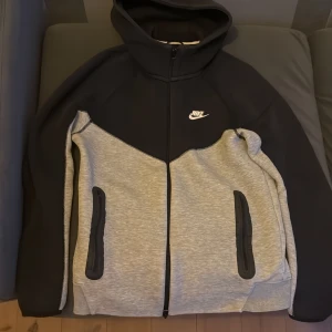Grå och svart nike tech - Säljer en snygg grå och svart huvtröja från Nike med dragkedja och fickor framtill. Perfekt för en avslappnad stil. Huvan ger extra komfort och värme. Den är lite sliten vid högra armbåge, och den har två knappt synbara rost fläckar på framsidan, sen tre små blå prickar på vänster arm 
