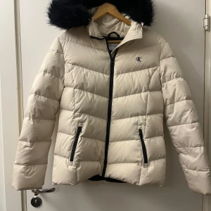 Vit dunjacka från Calvin Klein - Snygg vit dunjacka från Calvin Klein med svart fuskpäls på huvan. Jackan har dragkedja och två fickor med svarta detaljer. Perfekt för kyliga dagar med stil. 🖤