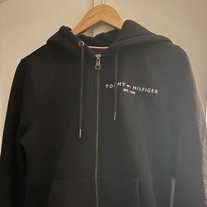 Svart hoodie från Tommy Hilfiger - Säljer en stilren svart hoodie från Tommy Hilfiger med dragkedja och justerbar huva. Den har märkets logga broderad på bröstet och är perfekt för en avslappnad look. Passar bra till både jeans och joggers.