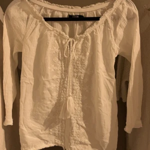 VIT BLUS - Superfin vit blus!!! Säljer då den är för liten för mig. Super fina detaljer med blomliknande mönster och snören med tofsar på som man kan knyta till önskad passform. Passar både off shoulder och som vanlig tröja. Lite kortare ärmar.