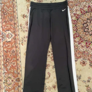 Svarta träningsbyxor från Nike - Snygga svarta träningsbyxor från Nike med vita sidopaneler. Perfekta för träning eller en avslappnad dag. Byxorna har en elastisk midja för extra komfort.