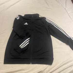 Svart träningsjacka från Adidas - Säljer en svart träningsjacka från Adidas med klassiska vita ränder längs ärmarna. Jackan har dragkedja framtill och en hög krage. Perfekt för sportiga aktiviteter eller som en stilren vardagsjacka.