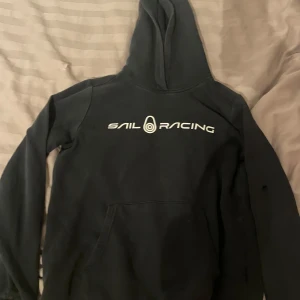 Svart hoodie från Sail Racing - Säljer en svart hoodie från Sail Racing med vit logga på bröstet. Tecken på användning som ses på bilden. 
