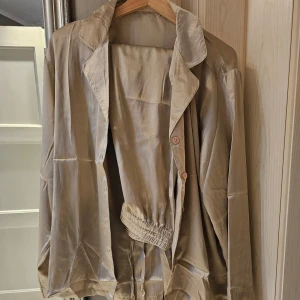 Beige sidenpyjamas/loungeset - Säljer en beige pyjamas/loungeset i siden med långärmad skjorta och matchande byxor. Skjortan har knappar framtill och en klassisk krage. Byxorna har en bekväm resår i midjan med knytband. Perfekt för en mysig kväll hemma. OANVÄNT