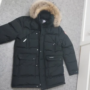 Svart dunjacka med päls från Canada Goose - Säljer en svart dunjacka från Canada Goose med pälsdetaljer på huvan. Jackan har långa ärmar och flera praktiska fickor med knappar och dragkedja. Perfekt för kalla vinterdagar.