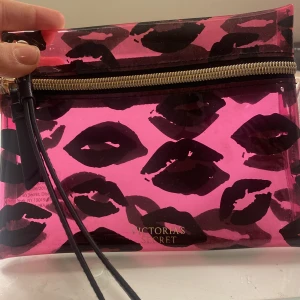 Transparent rosa kuvertväska från Victoria's Secret - Snygg transparent rosa kuvertväska från Victoria's Secret med svarta läppmönster. Väskan har en dragkedja i guld och en svart handledsrem. Perfekt för att hålla dina småsaker organiserade med stil.