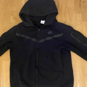 Svart hoodie från Nike - Säljer en svart hoodie från Nike med dragkedja och huva. Den har ett diskret Nike-logotyp på bröstet och en stilren design med svarta detaljer. Perfekt för en avslappnad stil.