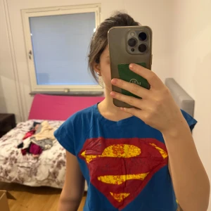Blå Superman t-shirt - Säljer en cool blå t-shirt med Superman-logga i rött och gult. T-shirten är croppad och har korta ärmar, perfekt för en avslappnad stil. Passar bra för alla superhjältefans! 💥