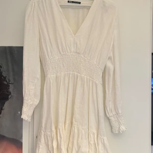 Linne klänning från Zara - en vit linne klänning från Zara med v-ringning och långa ärmar. Klänningen har en smockdetalj i midjan som ger en fin passform och volangdetaljer vid ärmslut och nederkant. Perfekt för vår och sommar!