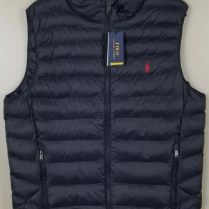 Svart dunväst från Polo Ralph Lauren - Snygg svart dunväst från Polo Ralph Lauren med dragkedja och broderad logga i rött. Perfekt för kyligare dagar och enkel att matcha med olika outfits.