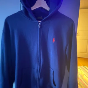 Blå hoodie från Polo Ralph Lauren - Säljer en snygg blå hoodie från Polo Ralph Lauren med dragkedja och det klassiska röda logotypen på bröstet. Perfekt för en avslappnad stil. Hoodien har en bekväm passform och är tillverkad i mjukt material. Inga skador✅⚡️