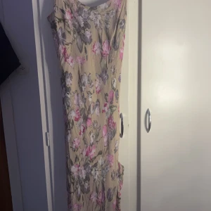 Blommig långklänning från Per Una - Säljer en vacker långklänning från Per Una med ett blommigt mönster i rosa, lila och grå toner på en beige bakgrund. Klänningen har smala axelband och en elegant passform. Perfekt för sommarens alla tillfällen! 🌸