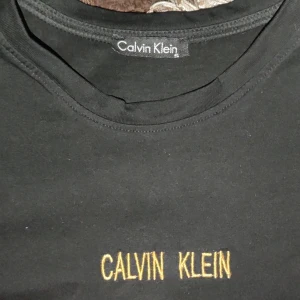 Svart t-shirt från Calvin Klein - Snygg svart t-shirt från Calvin Klein med guldfärgad logga på bröstet. Perfekt för en stilren look.