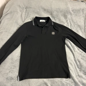Svart långärmad pikétröja från Stone island - Snygg svart långärmad pikétröja från Stone island med vit randdetalj på kragen. Sprillans ny då jack fick den för ett tag sen men den har bara legat i garderoben, fortfarandd lapparna kvar. Nypris 1349