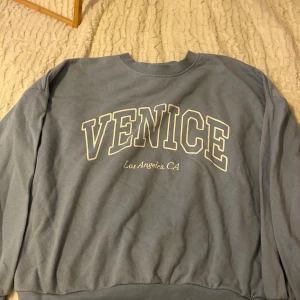 Blå sweatshirt från H&M - Säljer en blå sweatshirt från H&M med texten 'VENICE Los Angeles, CA' tryckt på framsidan. Tröjan har en rund halsringning och långa ärmar. Perfekt för en avslappnad stil.