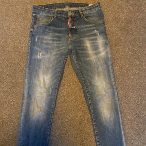 Blå jeans från DSquared2 - Snygga blå jeans från DSquared2 med slitna detaljer och en klassisk femficksdesign. De har en knappgylf och en röd logga på bakfickan. Inga skador✅