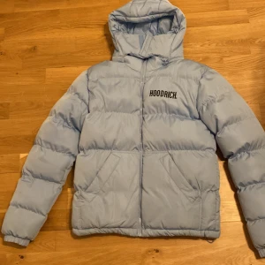 Ljusblå pufferjacka från Hoodrich - Säljer en snygg ljusblå pufferjacka från Hoodrich. Jackan har en huva och dragkedja framtill. Den är vadderad och har märkets logga både fram och bak. Perfekt för kyliga dagar!
