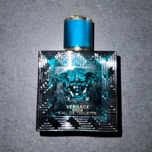 Versace Eros Eau de Toilette - Säljer en flaska Versace Eros Eau de Toilette. Flaskan är fyrkantig och har en djup turkos färg med ett elegant mönster och Versaces ikoniska Medusa-logga. Korken är också turkos med gulddetaljer. Perfekt för den som vill ha en doft med karaktär och stil.