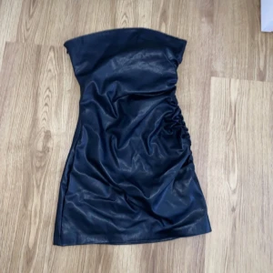 Svart axelbandslös klänning - Snygg svart axelbandslös klänning i syntetmaterial med rynkade detaljer på sidan. Perfekt för en kväll ute eller en speciell tillställning. Klänningen har en elegant och stilren design.