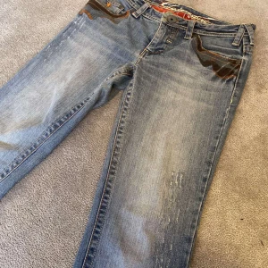 Blå jeans med unika detaljer - Snygga blå jeans från Roberto Cavalli med unika bruna detaljer på fickorna. De har en klassisk femficksdesign och en knappgylf. En av knapparnas design har ramlat av men funkar fortfarande. 