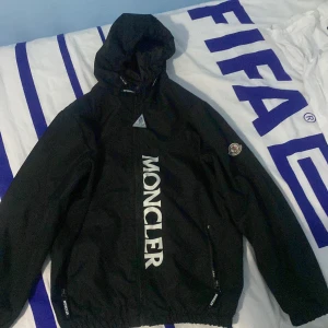 Svart vindjacka från Moncler - Säljer en stilren svart vindjacka från Moncler med stor logga på framsidan. Jackan har en praktisk huva och dragkedja framtill. Perfekt för blåsiga dagar!