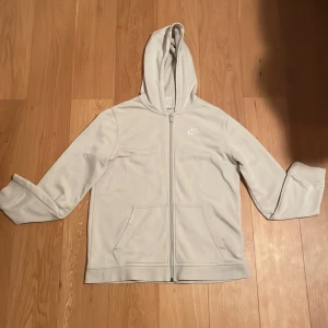 Vit/ beige hoodie från Nike - Säljer en stilren vit hoodie från Nike med dragkedja och huva. Har haft den i cirka 1 år men inte använt mycket.  Storlek 158-170cm  
