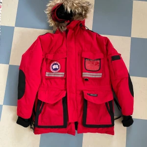 Röd vinterjacka Canada Goose -  varm o skön i jättefint skick. Kommer från ett förråd och är över 12 år gammal men i väldigt gott skick och sparsamt använd. Den har en liten svag blek fläck under ena armen annars i mycket gott skick. Xs men stor i storleken, funkar upp till M skulle jag säga. Tror 100% att den är äkta men vet inte ursprung,  därav priset. Till skillnad från kopior så är den tung, flera smarta fickor på insidan, tät i stygnen (inga lösa trådar), rejäla dragkedjor mm. 