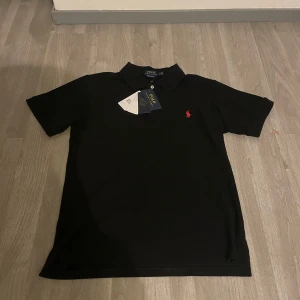 Svart pikétröja från Ralph Lauren - Snygg svart pikétröja från Ralph Lauren i custom slim fit. Tröjan har en klassisk krage och korta ärmar. Den är dekorerad med det ikoniska röda broderade logotypen på bröstet. Perfekt för en stilren look.