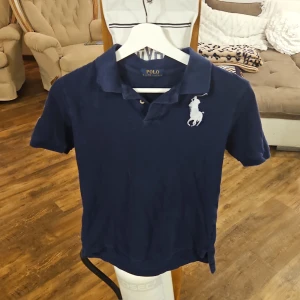 Mörkblå pikétröja från Ralph Lauren - Säljer en snygg mörkblå pikétröja från Ralph Lauren med korta ärmar. Tröjan har en stor vit logga på bröstet och siffran '3' på ärmen. Nypriss runt 700 storlek s