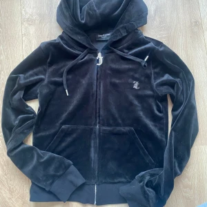 Svart velourjacka från Juicy Couture - Snygg svart velourjacka från Juicy Couture med dragkedja och huva. Jackan har ett stort 'Juicy' tryck i strass på ryggen och en liten logga på bröstet. Perfekt för en avslappnad och stilren look.
