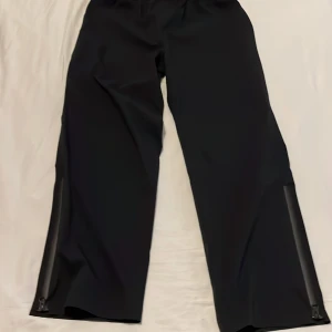 Svarta  golf/regnbyxor från DryJoys - Svarta regnbyxor från DryJoys med elastisk midja och dragkedjor vid bensluten. Tillverkade i polyester med HydroLite-teknologi för att hålla dig torr. Perfekta för regniga dagar.
