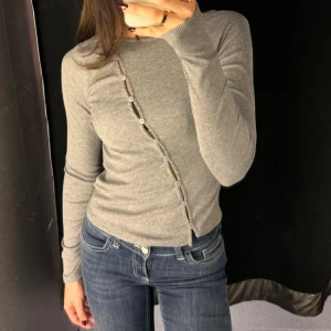 Grå ribbad tröja med knappar - Säljer en stilren grå ribbad tröja med unika knappar i en diagonal linje framtill. Tröjan har långa ärmar och en tight passform, perfekt för en trendig look.