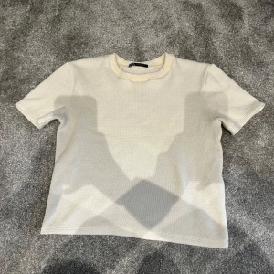 Beige kortärmad tröja från Zara - Säljer en stilren beige kortärmad tröja från Zara. Tröjan har en rund halsringning och är tillverkad i ett mjukt material som ger en bekväm passform.