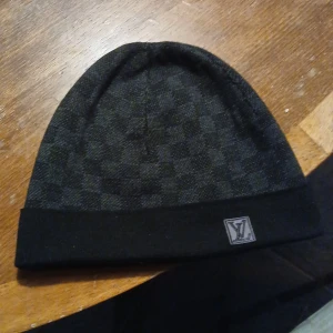 Svart rutig mössa från Louis Vuitton - Snygg svart mössa med diskret rutmönster från Louis Vuitton. Mössan har en uppvikt kant och en broderad logotyp framtill. Perfekt för att hålla sig varm med stil. Har tyvärr inte kvitto