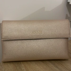 Beige axelväska från Guess - Snygg beige axelväska från Guess med kedjerem och magnetlås. Väskan har en stilren design med Guess-logga på framsidan och flera fack inuti för smidig organisering. Perfekt för att bära med sig det viktigaste med stil.