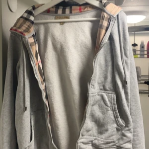 Burberry hoodie - Säljer en stilren grå hoodie från Burberry med dragkedja och klassiskt rutigt mönster på insidan av huvan. Tröjan har broderad logga på bröstet och praktiska fickor framtill. Perfekt för en avslappnad och trendig look. Den är i storlek L men passar M med. Säljer för att den är för liten i armarna 