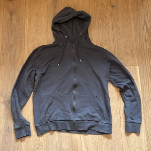 Grå hoodie med dragkedja - Säljer en stilren grå hoodie med dragkedja och justerbar huva. Perfekt för en avslappnad look. Tröjan har långa ärmar och praktiska fickor framtill. Köptes för 300kr nu säljs för 200kr