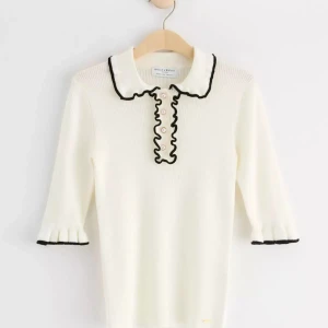 Vit ribbad topp med svarta detaljer - Säljer en stilren vit ribbad topp från Holly & White med svarta kanter och volangdetaljer framtill. Toppen har trekvartsärmar och en klassisk krage. Perfekt för en chic look!