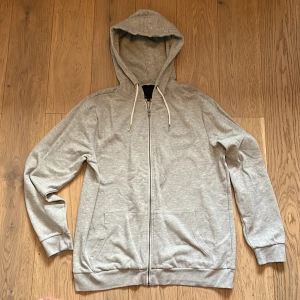 Grå hoodie med dragkedja - Säljer en stilren grå hoodie med dragkedja och fickor framtill. Perfekt för en avslappnad look. Den har långa ärmar och en justerbar huva med snören. Passar bra till både jeans och träningsbyxor.köpte för 300kr säljs för 200kr