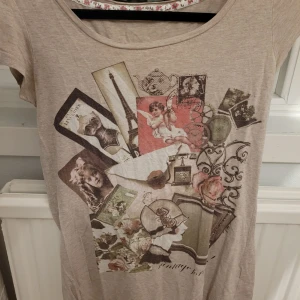 Beige t-shirt med vintage-tryck - Säljer en beige t-shirt med ett unikt vintage-inspirerat tryck på framsidan från Promod. T-shirten har korta ärmar och en rund halsringning. Perfekt för en avslappnad stil med en touch av retro. Står ingen storlek men skulle gissa S-M. Den har ett litet hål vid armhålan och syns knappt om man inte kollar noga