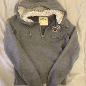 Grå hoodie från Hollister - Mysig grå hoodie från Hollister med mjukt foder i luvan och dragkedja framtill. Den har en broderad logga på bröstet och justerbara snören i luvan. Perfekt för kyliga dagar! Köpt från Sellpy, men väldigt bra skick och gosig💕