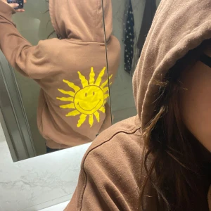 Brun zipup hoodie med tryck - Säljer en jätte mysig brun hoodie med dragkedja och ett gult soltryck på bröstet och ryggen. Tröjan har en luva med snören och långa ärmar. 