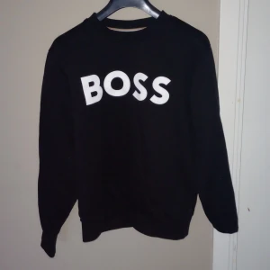 Svart tröja från BOSS - Säljer en stilren svart tröja från BOSS med vit text på framsidan. Tröjan har långa ärmar och en klassisk passform. Tröjan är i storlek s.