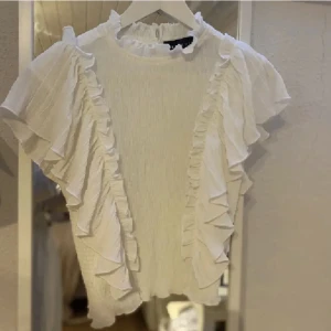 Vit blus från Never Denim - Säljer en elegant vit blus från Never Denim med volangdetaljer längs ärmarna och framsidan. Blusen har en hög krage och korta ärmar med volang. Perfekt för en stilren look.