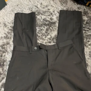 Svarta kostymbyxor från Dressmann - Snygga svarta kostymbyxor från Dressmann i slim fit. Byxorna har en klassisk design med knappar och dragkedja framtill. Perfekta för en stilren look.