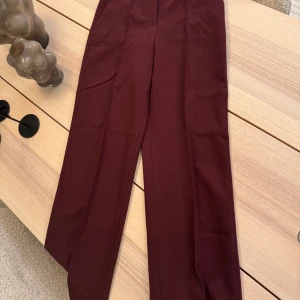 Vinröda kostymbyxor - Snygga vinröda kostymbyxor med hög midja och rak passform. Perfekta för en stilren look. Byxorna har en diskret dragkedja och knappar för en elegant finish. Sitter jätte fint i ninjan och är inte tajta som vissa byxor brukar vara. Lagom köpta i november 2024 bara en gång har jag använt dem.