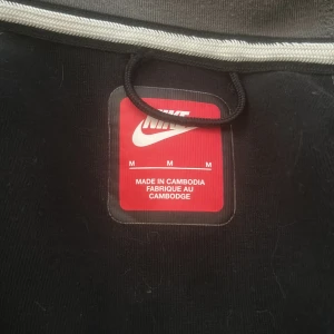 Grå och svart hoodie från Nike - Säljer en grå och svart hoodie från Nike med dragkedja och huva. Jackan har långa ärmar och två fickor framtill. Perfekt för en avslappnad stil.